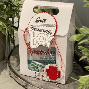 Könnte beinhalten: Eine weiße Geschenkbox mit einer rot-weißen Schleife. Die Box hat einen roten Kreis mit dem Text "Gute Besserung's Box" und einem Thermometer. Die Box ist gefüllt mit verschiedenen Artikeln, darunter eine Flasche Medizin und eine Packung Hustenbonbons.