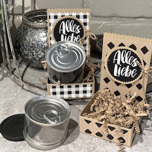 Könnte beinhalten: Zwei Geschenkboxen mit schwarz-weißem Karomuster und schwarz-braunen Rautenmustern. Jede Box enthält eine Dose mit einer durchsichtigen Plastikabdeckung und braunem Schredderpapier. Die Boxen haben einen schwarzen Kreis mit weißem Text, der "Alles Liebe" sagt.
