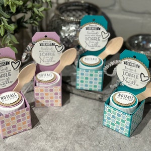 Könnte beinhalten: Vier kleine Geschenkboxen mit einem karierten Muster in Rosa, Türkis und Blau. Jede Box enthält ein kleines Glas mit einem braunen Deckel und einen Holzlöffel. Ein Schild mit dem Text "Wenn du denkst du kannst nicht mehr, dann löffle ein Nutella" ist an jeder Box befestigt.