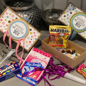 Könnte beinhalten: Eine braune Box mit einem pink-grünen Punkte-Muster und dem Text "Gute Laune Box" auf dem Deckel. Die Box ist gefüllt mit verschiedenen Kleinigkeiten, darunter Süßigkeiten, eine Seifenblasen-Puste und ein kleines Säckchen mit rosa Pulver.