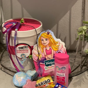 Könnte beinhalten: Ein rosa-weißer Eimer mit einem Meerjungfrauen-Design und dem Text "KIDS VERWÖHN PROGRAMM". Der Eimer ist gefüllt mit Badeprodukten, Süßigkeiten und Badebomben. Zu den Badeprodukten gehören eine rosa Flasche Shampoo und eine blaue Flasche Schaumbad. Die Süßigkeiten umfassen eine Tüte Gummibärchen und eine Schachtel Smarties. Die Badebomben sind in Form eines Sterns und einer Meerjungfrau.