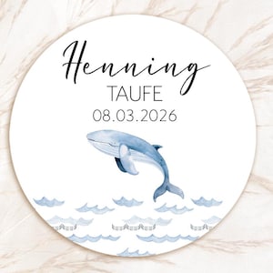 Op de afbeelding: Een ronde witte sticker met de naam "Henning" en het woord "TAUFE" in zwart schrift. Een blauwe walvisillustratie is afgebeeld boven blauwe golfafbeeldingen. De datum "08.03.2026" staat eronder.