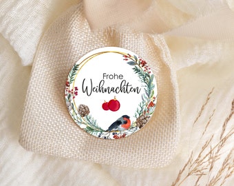 May include: A round white Christmas ornament with a gold border and a wreath of greenery, red berries, and a small bird. The ornament says "Frohe Weihnachten" in black text.