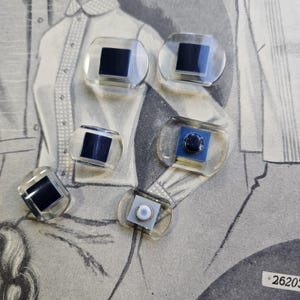 May include: A collection of six clear, oval-shaped cufflinks with a dark square or circular center. Some cufflinks have a black square, while others feature a blue square with a dark button or a white button.