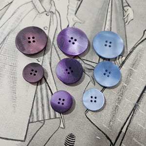 Peut inclure: Une collection de boutons ronds de différentes tailles et nuances de violet et de bleu. Les boutons ont quatre trous pour la couture et sont disposés sur une illustration de mode vintage. Les boutons mesurent entre 1,3 cm et 2,5 cm de diamètre.