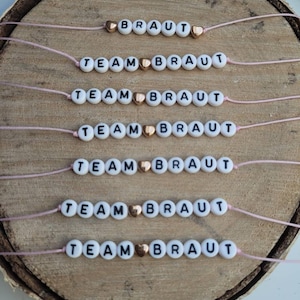 Könnte beinhalten: Sieben rosafarbene Armbänder aus Schnur mit weißen Perlen, die "TEAM BRAUT" buchstabieren und einem goldenen Herz-Anhänger.