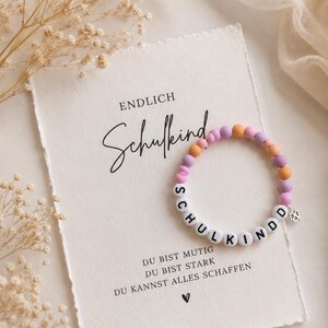 Könnte beinhalten: Ein Perlenarmband mit abwechselnden matten violetten, rosafarbenen und orangefarbenen Kugeln, weißen Buchstabenperlen mit der Aufschrift SCHULKIND und einem kleinen silbernen Kleeblatt-Anhänger, platziert auf einer Karte mit dem Text ENDLICH Schulkind DU BIST MUTIG DU BIST STARK DU KANNST ALLES SCHAFFEN.