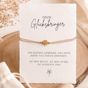 Könnte beinhalten: Ein Armband mit einem goldfarbenen vierblättrigen Kleeblatt an einem dünnen beigen Band, präsentiert auf einer weißen Karte mit dem Text: DEIN Glücksbringer, EIN KLEINES ARMBAND, DAS DICH JEDEN TAG DARAN ERINNERT: DU BIST MUTIG. DU BIST STARK. DU SCHAFFST DAS!