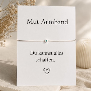 Könnte beinhalten: Ein dünnes beiges Schnurarmband mit einer einzelnen weißen Perle, die ein kleines türkisfarbenes Herz zeigt, präsentiert auf einer weißen Karte mit dem Text Mut Armband, Du kannst alles schaffen und einer schwarzen Herz-Strichzeichnung.