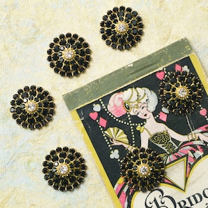 May include: Six black rhinestone flower-shaped embellishments with gold-tone metal settings. Each has a clear rhinestone center. The embellishments are scattered on a vintage playing card illustration.