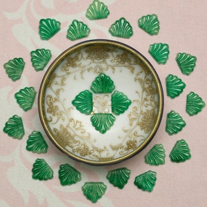 May include: A white and gold decorative bowl with a dark blue rim, surrounded by green, shell-shaped embellishments. The bowl features a floral design and a central green, four-leaf clover pattern. The background is a light pink fabric.