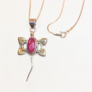 Puede incluir: Un collar de plata con un colgante que presenta una piedra preciosa roja engastada en un marco de oro y plata. El colgante tiene la forma de una libélula con una cola larga.