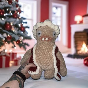 Op de afbeelding: Een bruin pluche wezen met een rood vest met witte rand en een witte hoofdbedekking. De handgemaakte pop heeft een grillig ontwerp met grote ogen en twee voortanden. De achtergrond omvat een kerstboom en een open haard.