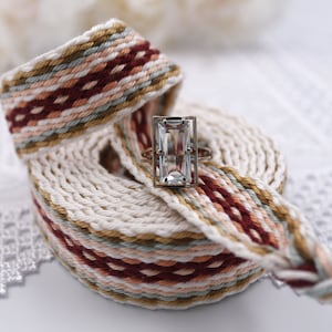 May include: A white, brown, and red woven band with a clear, rectangular gemstone ring on top.