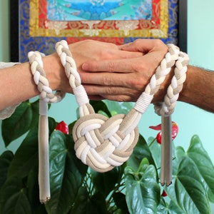 May include: Two people holding hands with a white and beige braided rope tied around their wrists. The rope is knotted into a heart shape in the center.