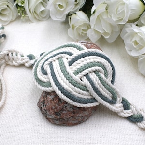 May include: A decorative nautical knot made of white, green, and gray rope. The knot is displayed on a small, textured rock, with a braided rope extending from it. White flowers are in the background.