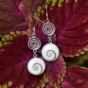 May include: A pair of silver earrings with a white shell pendant and a silver spiral design. The earrings are on a red and green leaf background.