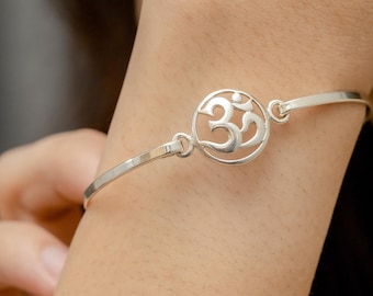 May include: A silver bangle bracelet with an openwork Om symbol charm. The bracelet is on a person's wrist.