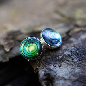 Puede incluir: Un par de pendientes de plata con incrustaciones de abulón iridiscente verde y azul. Los pendientes son redondos y tienen un acabado liso y pulido.