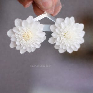 May include: Two white flower earrings with a light yellow center are held up by a clear plastic frame. The earrings are in the shape of a daisy. The background is a blurred gray.
