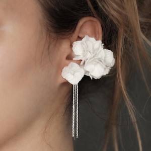 May include: A white floral ear cuff featuring layered flower petals and two delicate silver-toned chains that hang downward. The accessory is positioned along the upper cartilage of the ear, creating a layered botanical appearance with metallic accents.