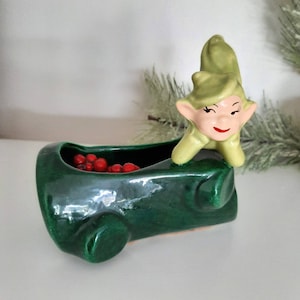 May include: A green ceramic planter shaped like a log with a smiling elf figurine. The planter is filled with red berries. The elf has a green hat and outfit with a pink face and red lips. A festive holiday decoration.