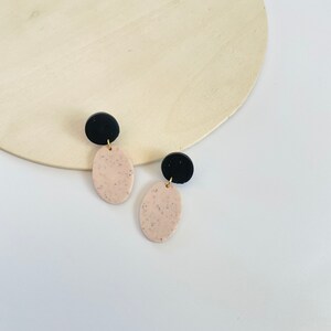 May include: A pair of dangle earrings with black and speckled beige oval-shaped charms. The earrings have a minimalist design.
