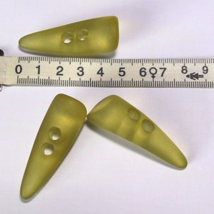 Könnte beinhalten: Drei gelbe Plastikknöpfe mit jeweils zwei Löchern. Die Knöpfe sind spitz oval geformt und etwa 2,5 cm lang.