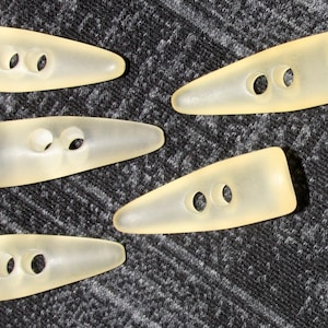 Peut inclure: Cinq boutons en plastique translucides de couleur jaune clair avec deux trous chacun. Les boutons ont la forme d'un ovale long et étroit.