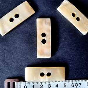 Op de afbeelding: Vier rechthoekige parelmoer knopen met elk twee gaten. De knopen zijn licht crème van kleur en hebben een licht iriserende glans.