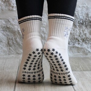 May include: Cream-coloured socks with black star patterns on the soles and ankles. The socks feature a ribbed design, two black stripes at the top, and a snowflake design. The words "MOON & GHOST" are printed on the soles.