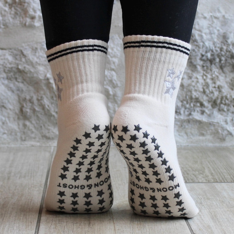May include: Cream-coloured socks with black star patterns on the soles and ankles. The socks feature a ribbed design, two black stripes at the top, and a snowflake design. The words "MOON & GHOST" are printed on the soles.