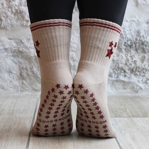 May include: Beige socks with maroon star patterns and stripes. The soles are covered in maroon stars and have the text "MOON & GHOSTS". The socks have a ribbed texture.