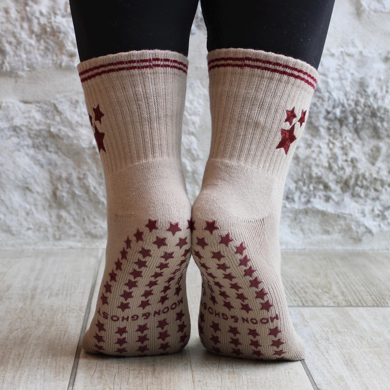 May include: Beige socks with maroon star patterns and stripes. The soles are covered in maroon stars and have the text "MOON & GHOSTS". The socks have a ribbed texture.