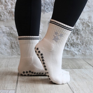 May include: Cream-coloured socks with black star grips on the soles and a black and white striped cuff. The socks feature a grey snowflake design on the side. The socks are ribbed and appear to be made of a soft material.