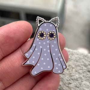 May include: A silver enamel pin of a ghost wearing daisy-patterned sunglasses. The ghost is wearing a lavender dress with white daisy accents.