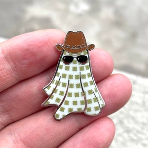 May include: A novelty enamel pin featuring a ghost wearing a brown cowboy hat and sunglasses. The ghost's body is white with a green checkered pattern, and the pin has a silver border. This pin is a fun accessory.