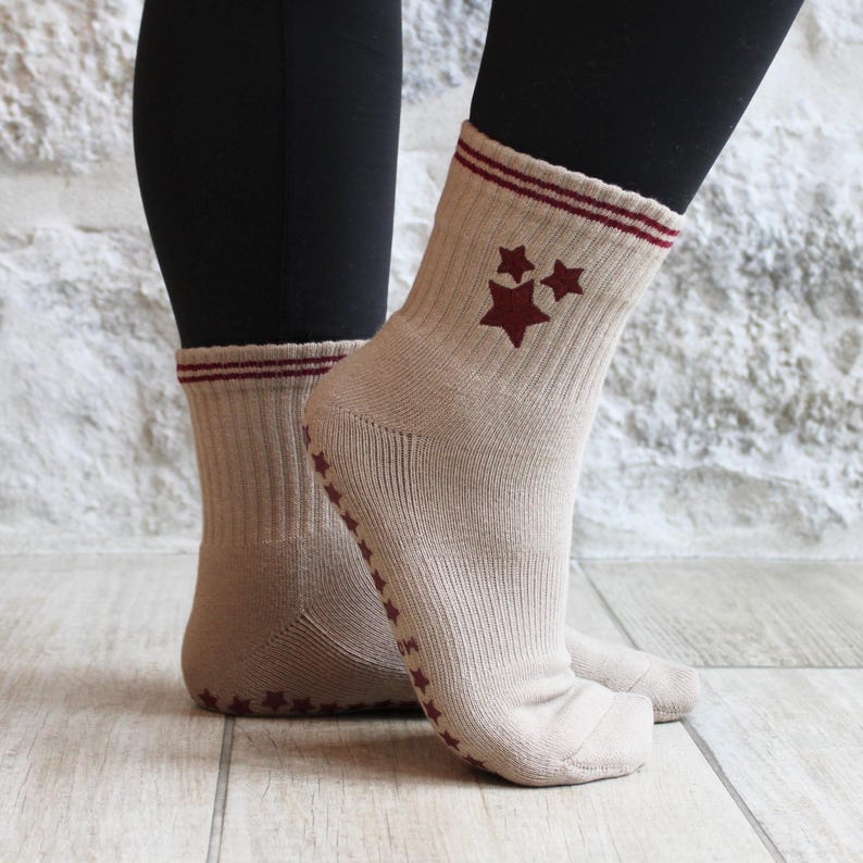 May include: Beige socks featuring maroon star patterns and maroon stripes at the top. The socks have a ribbed texture and non-slip star grips on the soles. The socks are worn with black leggings.