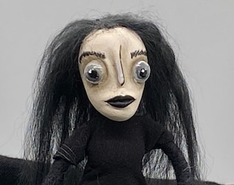 May include: A handmade doll with long black hair, large white eyes, and a black dress. The doll has a pale face with a painted-on nose and mouth.