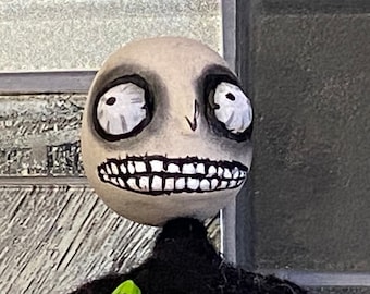 May include: A white doll head with black painted features, including large eyes, a wide smile with teeth, and a small nose. The doll's head is made of a textured material and has a slightly creepy appearance.