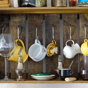 Könnte beinhalten: Ein Holzregal mit Haken, an denen verschiedene Tassen, ein Weinglas und eine Flasche hängen. Das Regal ist gelb gestrichen und hat einen rustikalen Look. Es gibt einen kleinen Teller mit einem grünen Objekt darauf und eine kleine schwarze Tasse auf dem Regal.