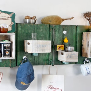 Könnte beinhalten: Ein grünes Holzregal an der Wand mit mehreren daran befestigten Holzkisten. Die Kisten sind mit verschiedenen Gegenständen gefüllt, darunter eine Kaffeedose, eine Glasflasche, eine Tasse und ein Hut. Ein Schild mit der Aufschrift "Caffe Florian" hängt an dem Regal.