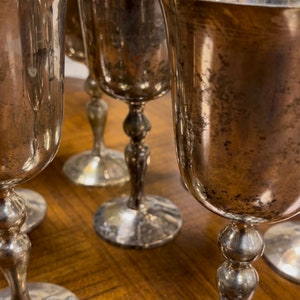 Könnte beinhalten: Ein Set aus sechs silbernen Kelchen mit Vintage-Look. Die Kelche haben einen runden Boden und einen hohen, schlanken Stiel. Die Kelche sind aus glänzendem Metall gefertigt und haben eine leicht angelaufene Optik.