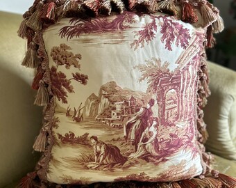 Op de afbeelding: Een vierkant kussen met een crèmekleurige achtergrond en een bruin en rood toile-patroon met een pastorale scène. Het kussen heeft een bruin en rood franje-bies.