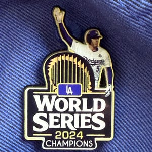 May include: A commemorative pin featuring a baseball player in a Dodgers uniform, celebrating the 2024 World Series Champions. The pin has a black and gold border with the words "LA WORLD SERIES 2024 CHAMPIONS". The background is a deep blue.