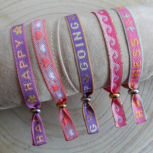 May include: Four colourful woven bracelets with different messages: "Happy", "Keep Going", "Fitness", and "Love". The bracelets are pink, purple, and green with gold lettering.
