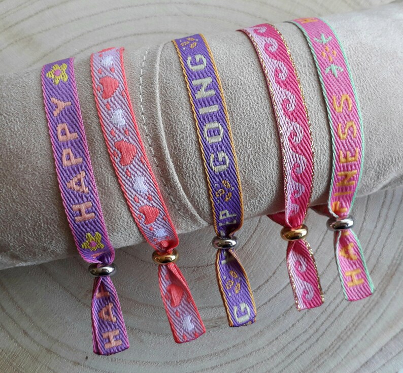 May include: Four colourful woven bracelets with different messages: "Happy", "Keep Going", "Fitness", and "Love". The bracelets are pink, purple, and green with gold lettering.