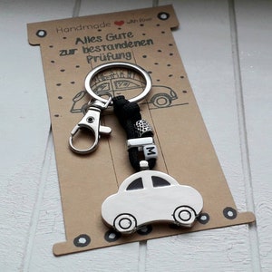 Könnte beinhalten: Ein silberner Auto-Schlüsselanhänger mit schwarzem Band und einem silbernen Anhänger. Der Schlüsselanhänger ist an einer Karte mit dem Text "Handmade with love Alles Gute zur bestandenen Prüfung" befestigt.