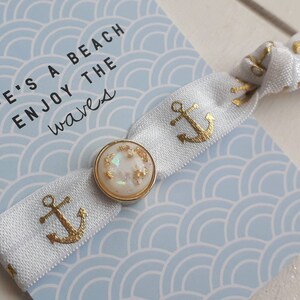 Könnte beinhalten: Ein weißes Gummiband für Haare mit goldfarbenem Ankerdruck und einem goldenen Knopf mit schimmerndem Glitzer. Das Haargummi befindet sich auf einem blau-weißen Hintergrund mit dem Text "Life's a beach enjoy the waves".