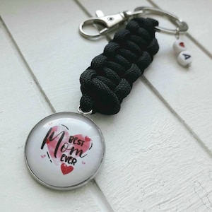 Könnte beinhalten: Schwarzer Paracord-Schlüsselanhänger mit einem silbernen Schlüsselring und einem runden Glasanhänger mit dem Text "Best Mom Ever" und einem roten Herzdesign. Der Anhänger ist mit einem silbernen Ring am Schlüsselanhänger befestigt.